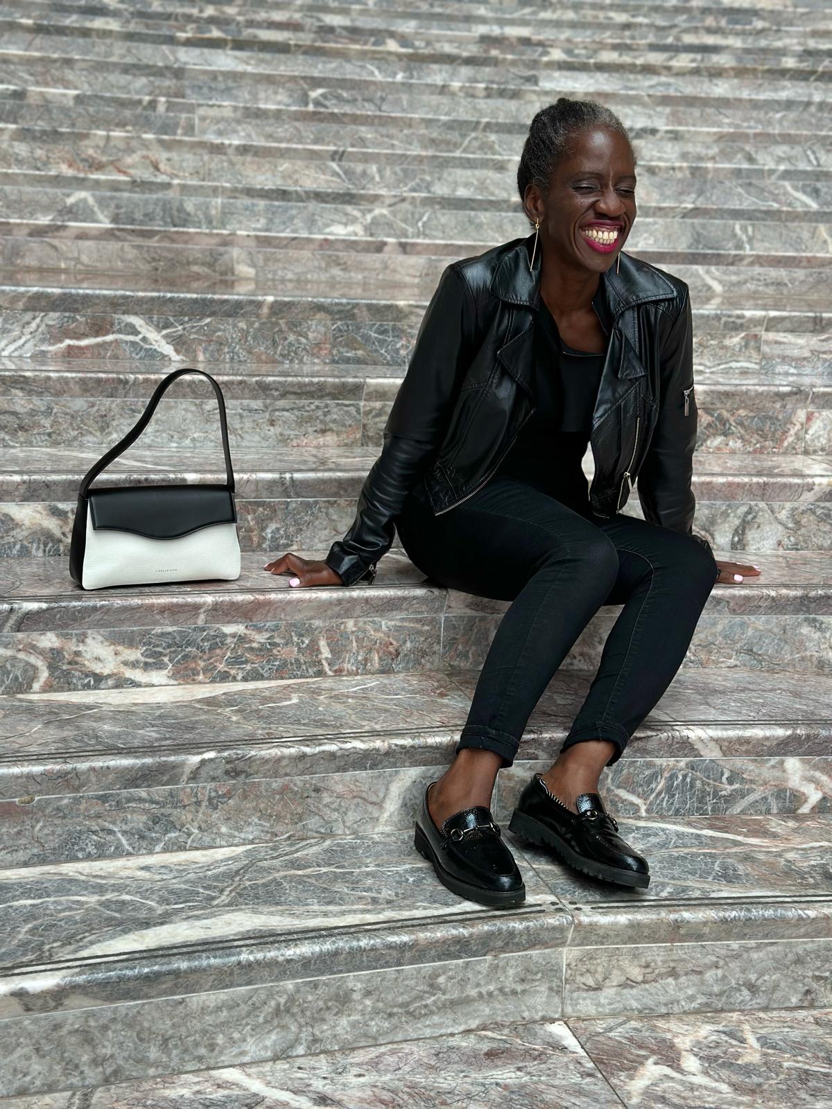 Finda smiling with her eyes closed, sitting on steps next to a designer handbag.